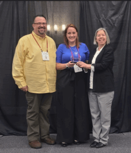 Three people stand together smiling in front of a black curtain. The woman in the center holds a glass award, while the man on the left and woman on the right stand beside her. All are wearing name badges.